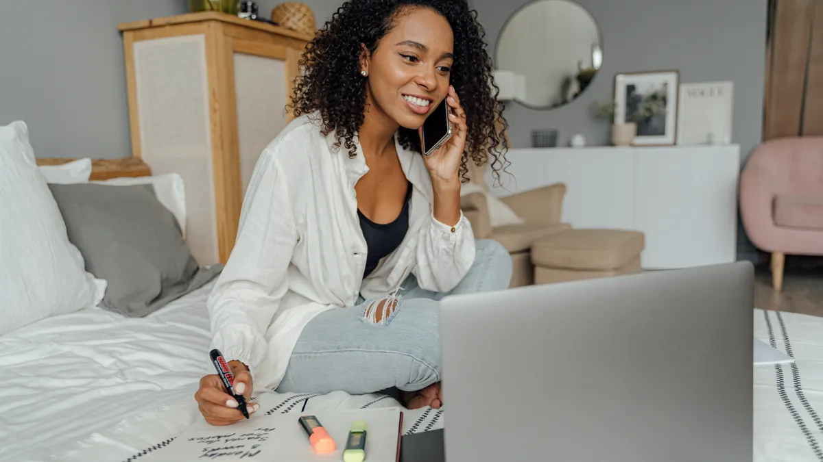 A feminine-presenting person types on a laptop and speaks on a phone