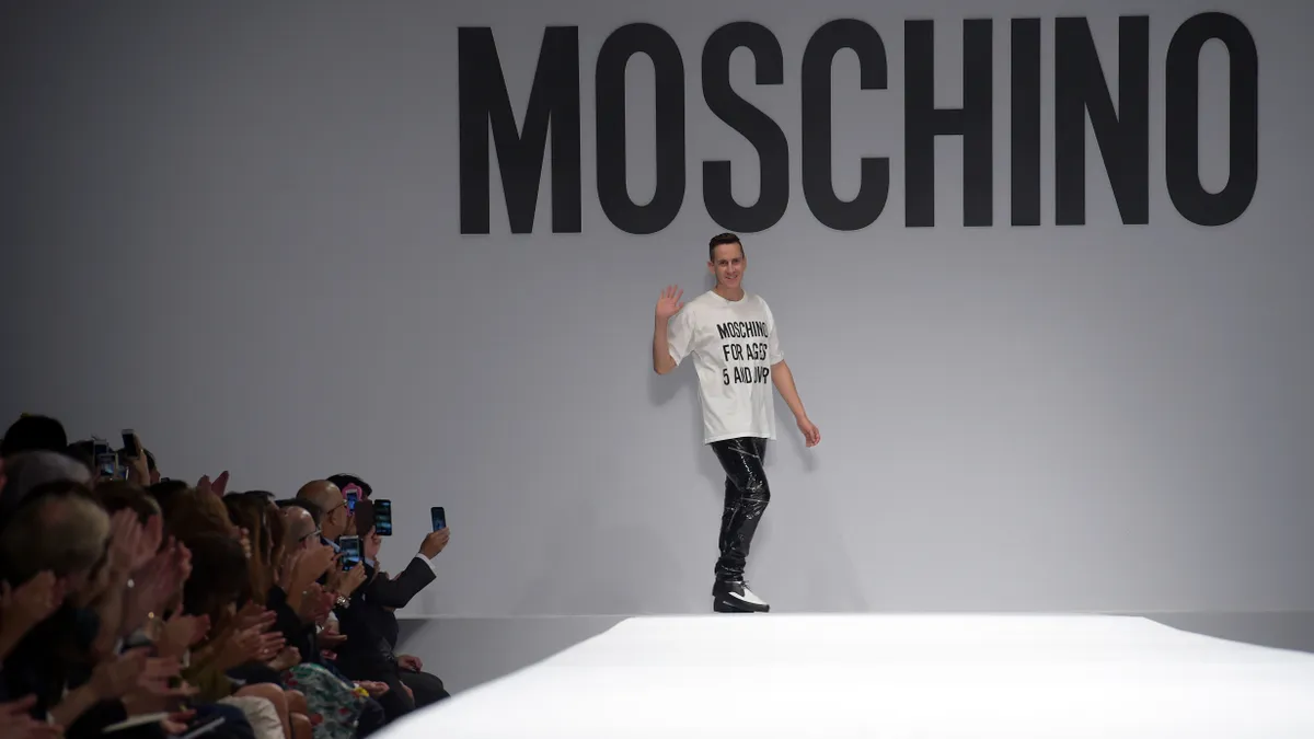 At the back of a runway, Jeremy Scott, wearing a white, t-shirt waves