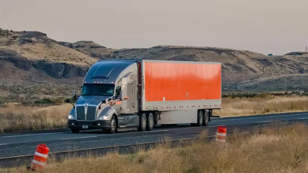 A Schneider National semi-truck traveling on a highway with rolling hillsides in the background.