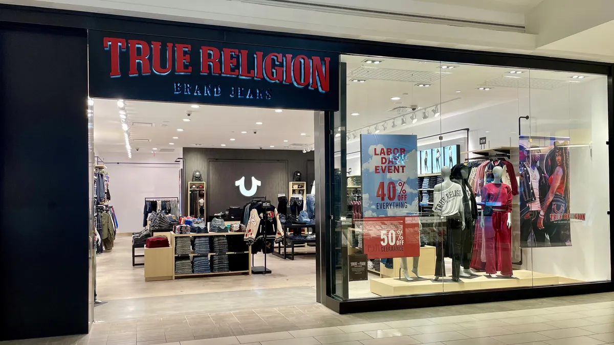 Exterior view shows a True Religion jeanswear store inside a mall with discount signs on the windows.