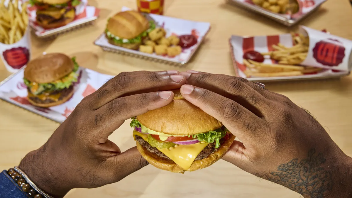 A pair of hands holding a burger over a table set with other burgers.