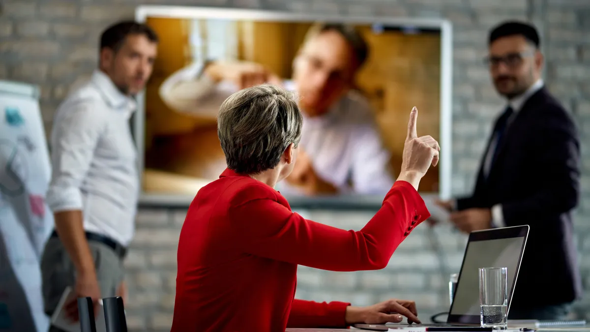 An executive wearing a bright jacket raises her hand in a meeting