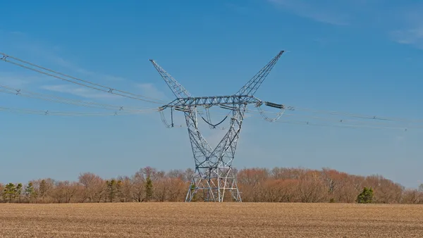 A transmission lIne pylon sits in the middle of a agricultural field with dry stubble with leafless trees in the background.