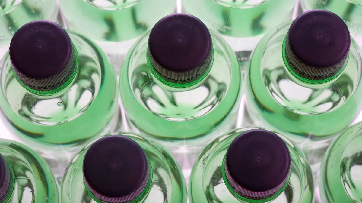 Overhead view of rows of green plastic sparkling water bottles with purple screw caps, arranged in a symmetrical grid pattern and illuminated from below, creating a vibrant, glowing effect.