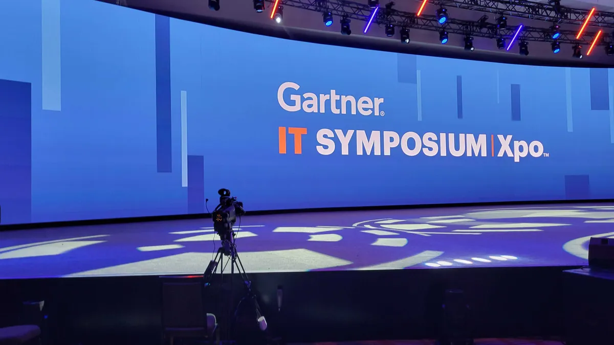 An empty stage lit with multicolored lights with a camera and a logo for Gartner IT Symposium/Xpo shown.