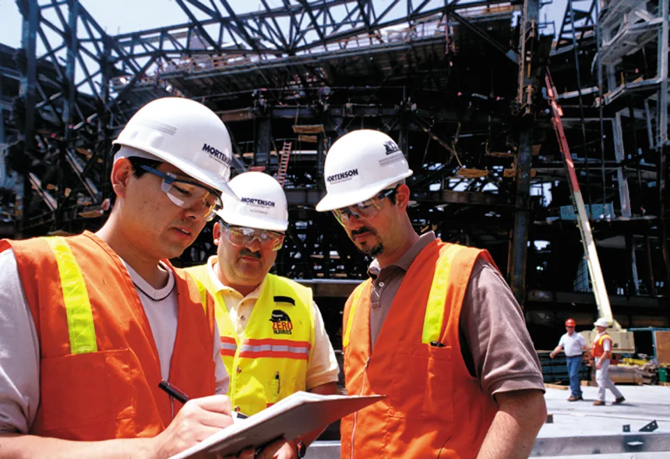 A group of people with construction equipment look at a clipboard at a job site.