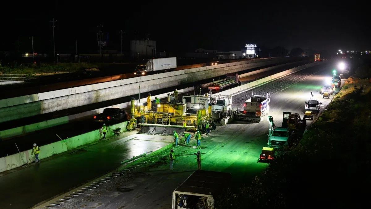 A construction crew in safety gear works on a highway project at night. Heavy machinery is visible in the illuminated area.