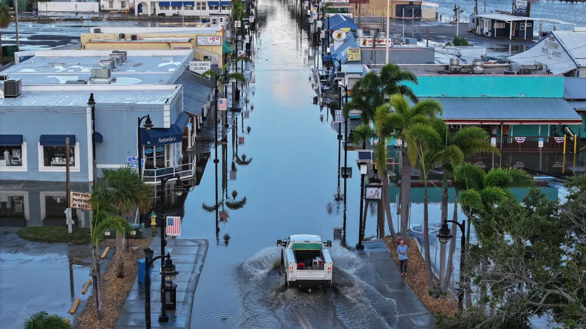 Aerial shot of flooded street lined by palm trees and houses. A truck drives down the flooded street.