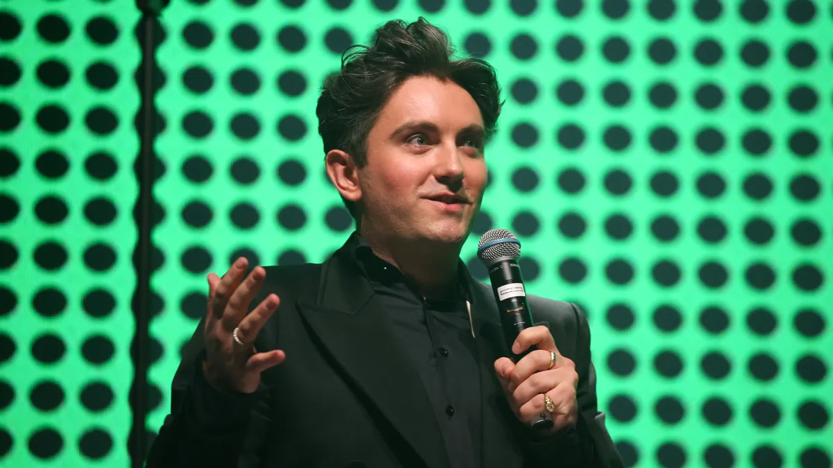 Man stands in front of green backdrop with black polka dots speaking into a microphone and gesturing with one arm.