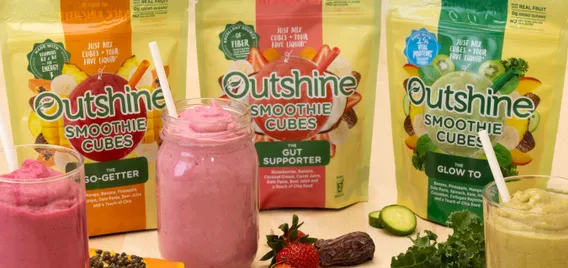 Jars containing smoothies made from Outshine Smoothie Cubes sit on a butcher block table, with the pouches that contain the cubes behind them. Fruits and vegetables are on the table around the jars.