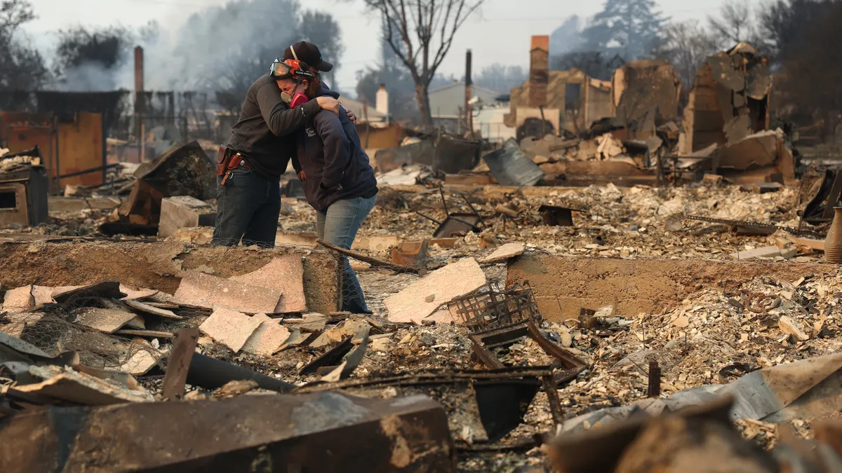 Two people embrace surrounded by charred buildings and rubble.