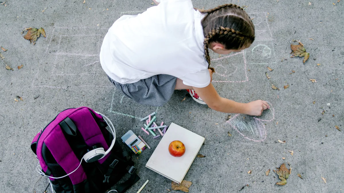 A young student uses chalk to draw on school asphalt. Next to them is a backpack and an apple sitting on a notebook.