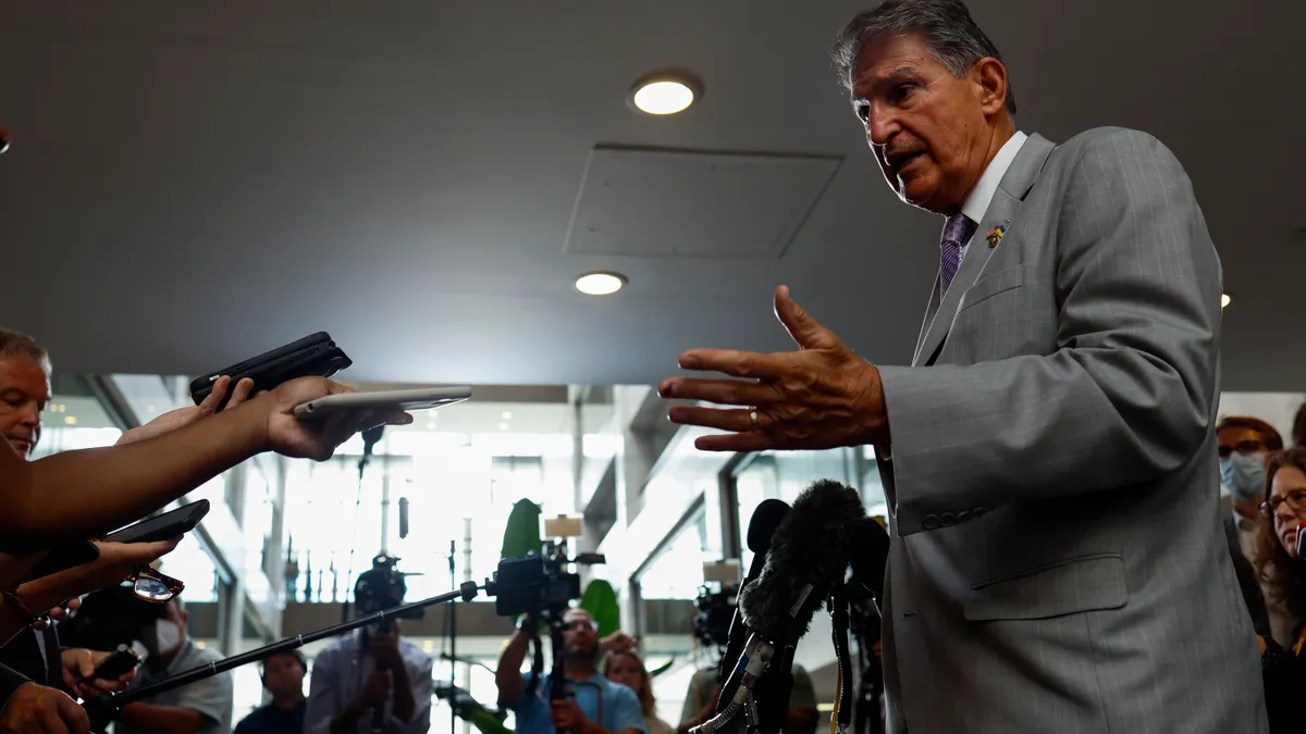 Man in suit talking to reporters, with microphones and phones in front of him