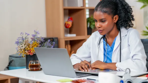 A focused doctor in a white coat, with a stethoscope, works on a laptop at a desk.