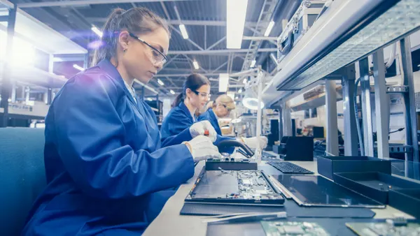 Electronics factory workers in blue work coats and protective glasses assembling a motherboard with a screwdriver.