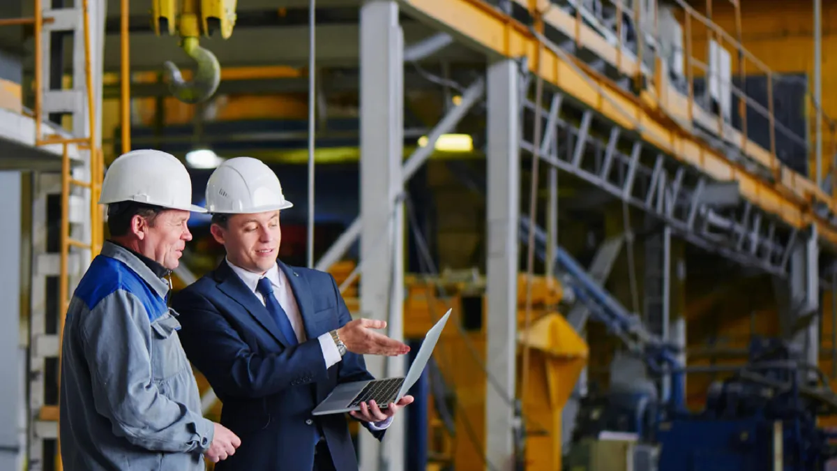 The man in the suit and the helmet holds the portable computer and shows up on the screen to the worker in overalls in an industrial building