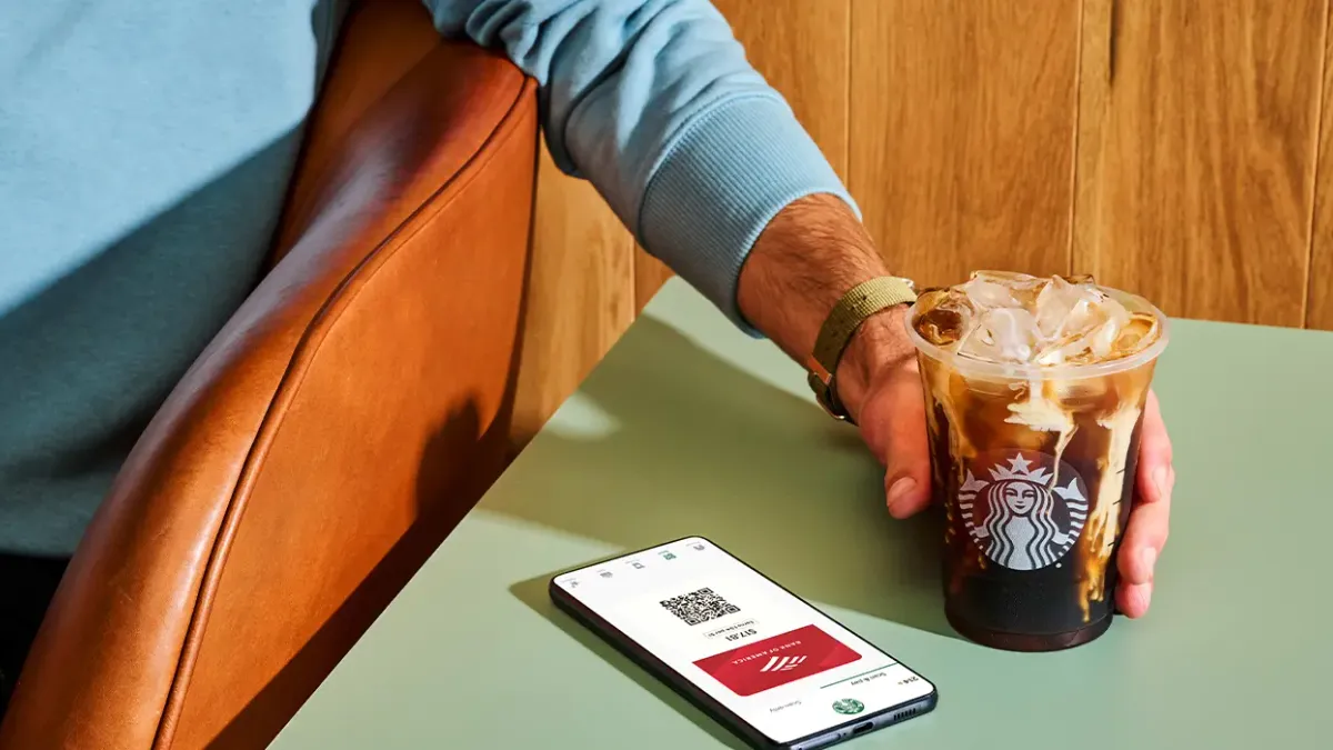 A man holds a starbucks drink next to a phone with a Bank of America card onscreen.