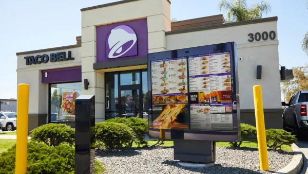 An image of a drive-thru digital menu board with a Taco Bell restaurant with white and purple paint in the background.