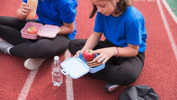 Two students sit together on a running track outside while eating from their lunch boxes.