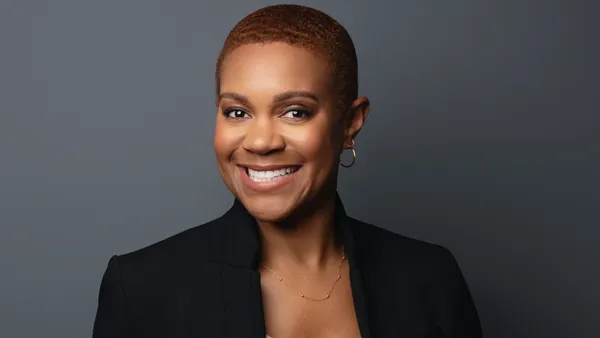 A person in a black suit jacket and short cropped hair smiles at the camera in a corporate headshot.