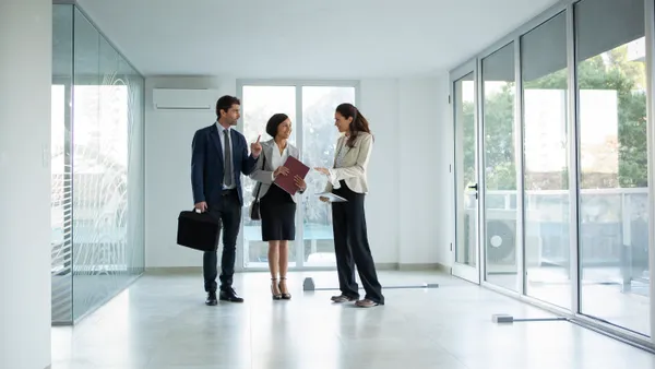 A wide shot of a female real estate agent talking with two businesspersons, while standing in empty office during daytime.