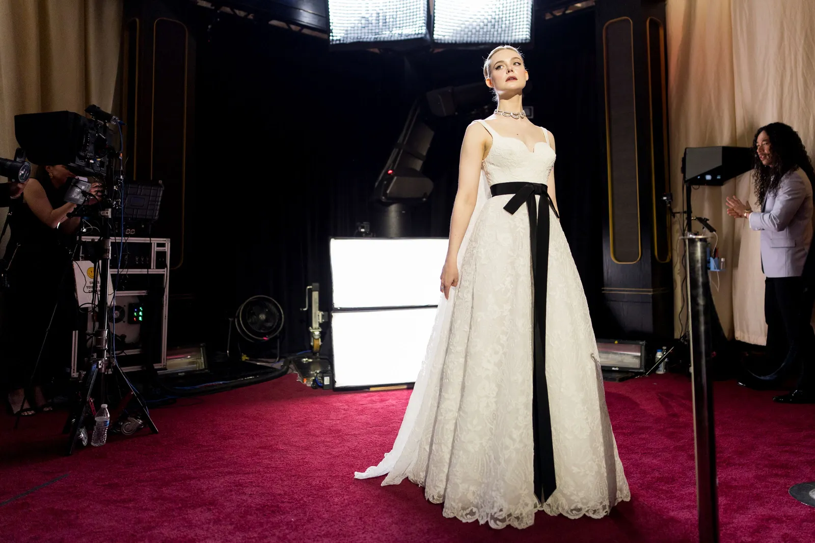 A woman in a long sleeveless white dress with a black bow belt stands on a red carpet as photographers aim lights at her.