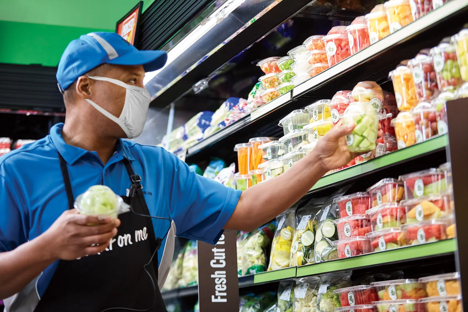 A worker wearing a face mask, cap and apron places a package of precut fruit on a grocery store shelf.