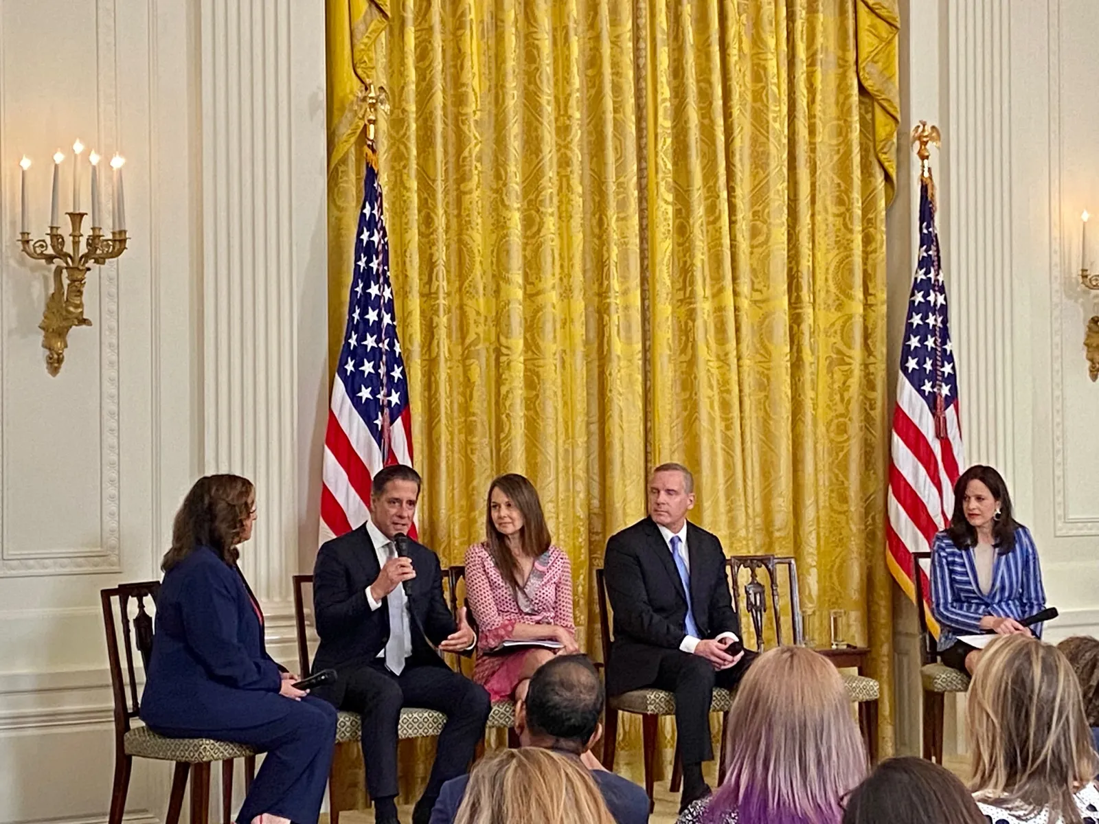 Five adults are sitting in chairs on a stage in front of a yellow curtain. One adult has a microphone to their face. On the stage are two flagpoles with American flags.