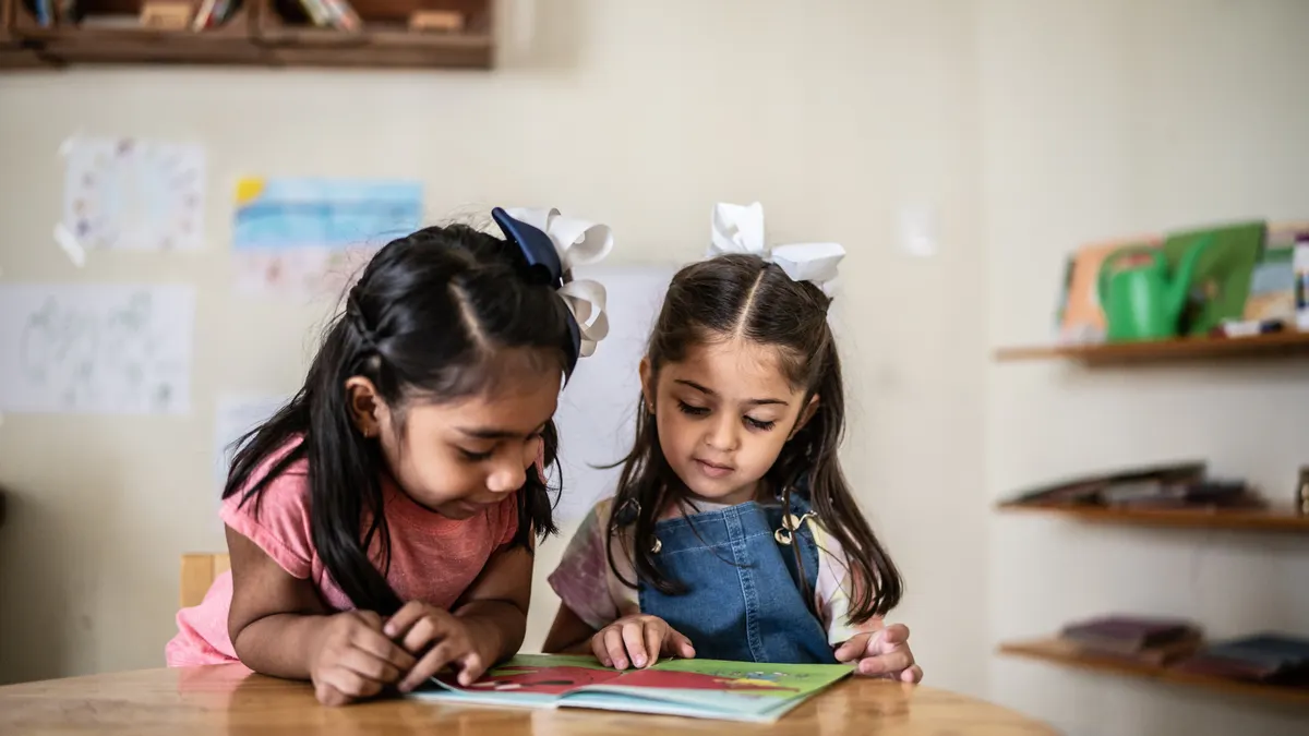Two young students are in a classroom. They are sitting at a desk and looking at a book together.