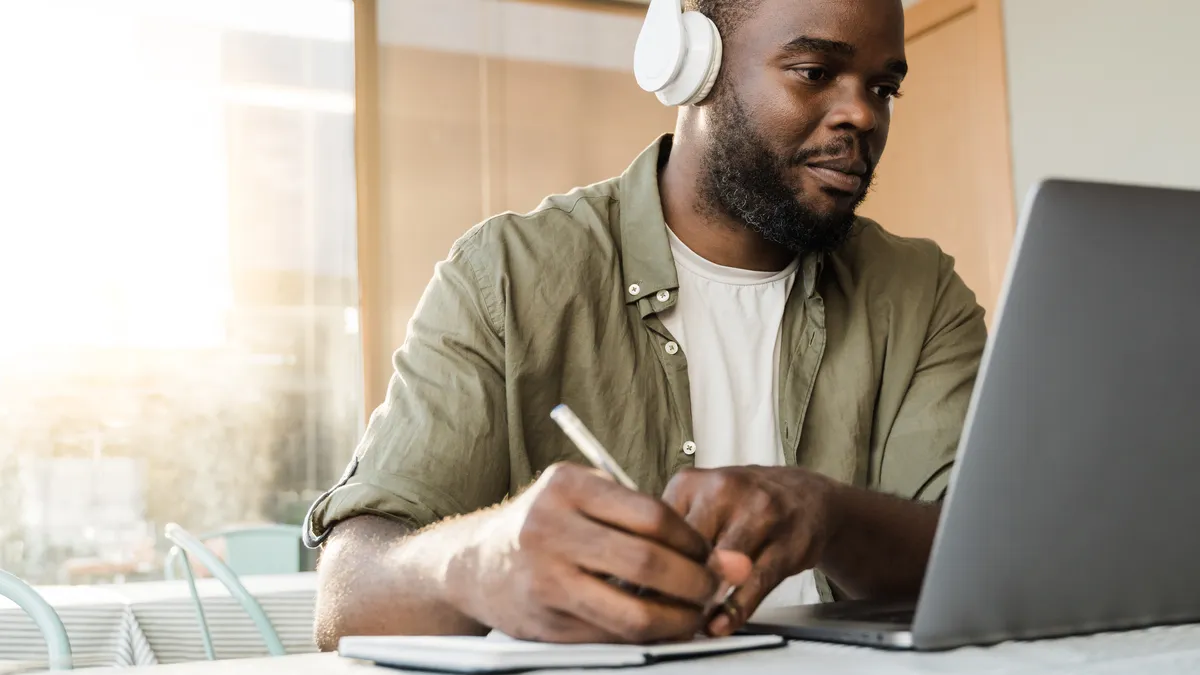 A man wearing headphones looks at a laptop and writes in a notepad.