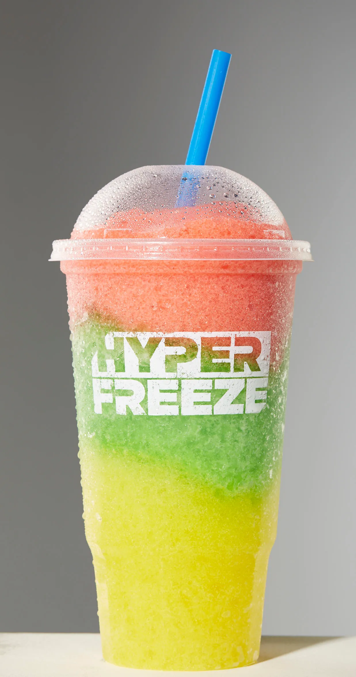 A photo of a frozen beverage in red, green and yellow layers and a blue straw in a clear plastic cup. The cup says &quot;Hyperfreeze.&quot;