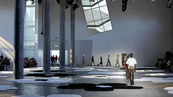 A wide shot of models walking in a line down a runway in a well-lit, cavernous room.