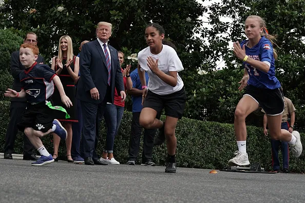 Then-President Donald Trump is pictured outside watching three children run in front of him. Other people are on the sidelines.