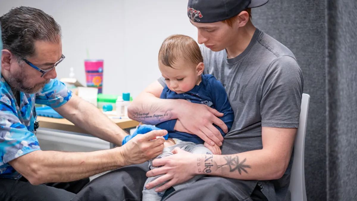 A young child gets a shot from a medical professional while another adult holds the child in their lap.