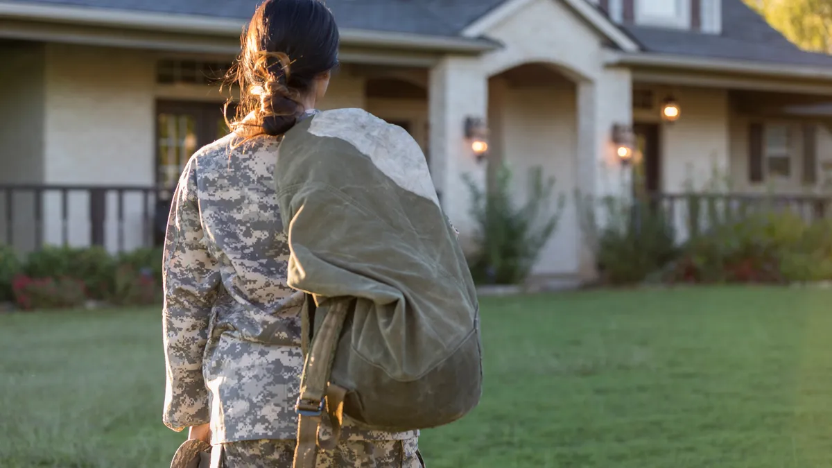 Person in military uniform and backpack with their back towards the camera looks at their home.