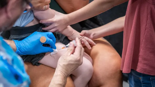 A syringe is being inserted into the leg of a child. A gloved hand holds a Baid-Aid. Another pair of hands seems to hold the arms of the child.
