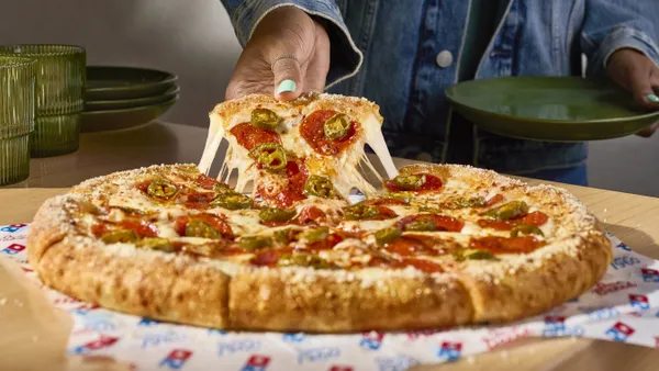 A hand lifts a slice of stuffed crust pizza topped with pepperoni and peppers. The slice trails strings of melted mozzarella.