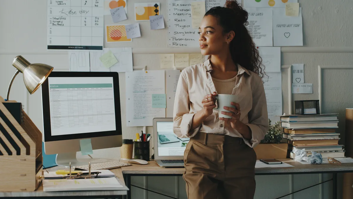 Young businessperson stands and looks contemplative while holding a cup of coffee in a home office