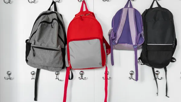 Four different colored backpacks are hanging on hooks on a wall