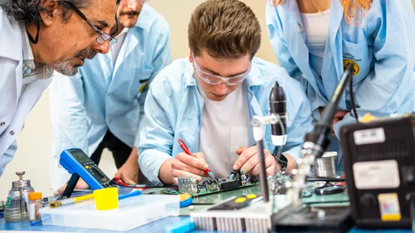 A student works on a robotics project while other students and a teacher watches.