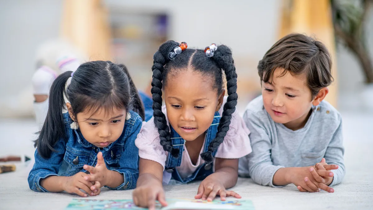 Three young children read the same book side by side while laying on the ground.