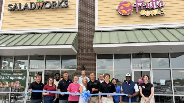 A group of people stand behind a blue ribbon and in front of a store that has two signs saying Saladworks and Frutta Bowls.