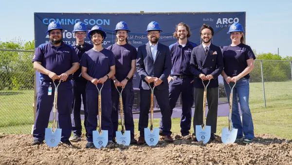 Eight people, some with hard hats and shovels, stand outside on a dirt site in front of a blue sign that says "coming soon."