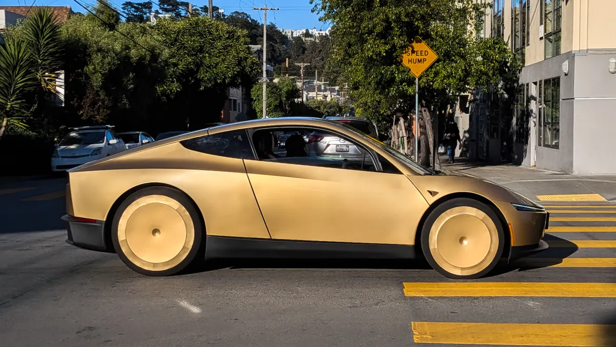 A sleek 2-door bronze color vehicle crosses an intersection with trees and buildings in the background.