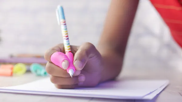A close up of a person's hands and fingers show the use of a pencil grip while writing in a notebook.