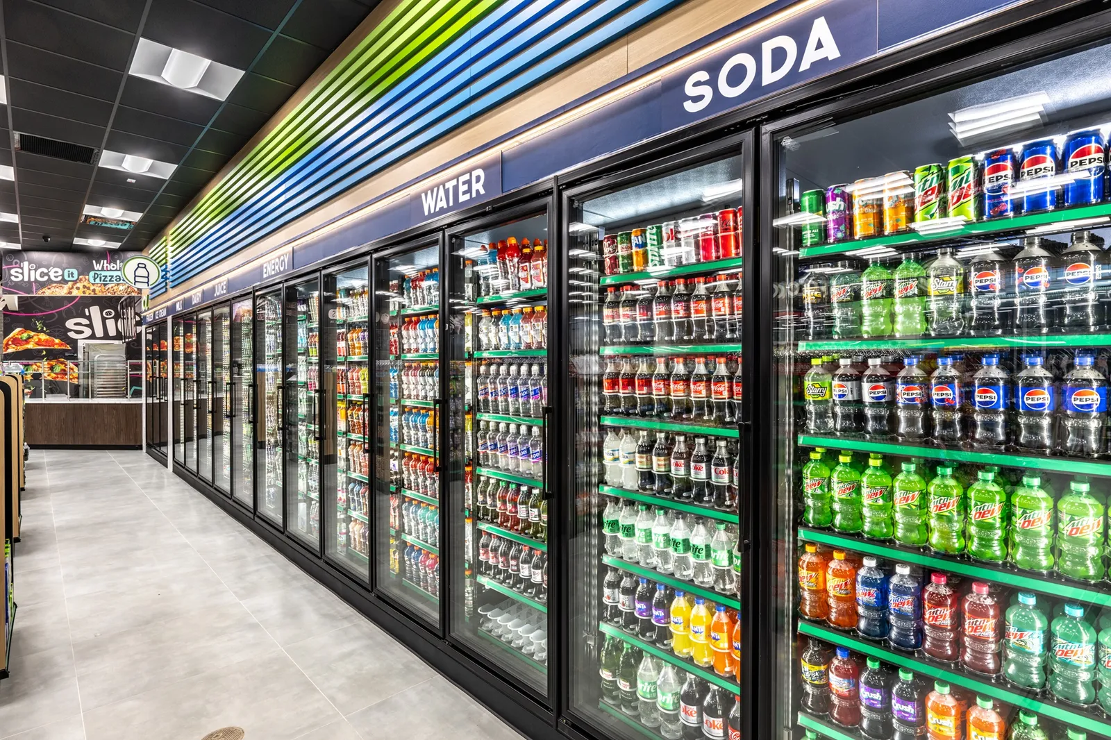 A photo of the interior of a store, showing a row of beverage coolers. Signs over the cooler doors say soda, water, energy, juice, and others. A sign on the wall in the back shows pizza and says Slice It. The store is decorated in greens and blues along with wood tones.