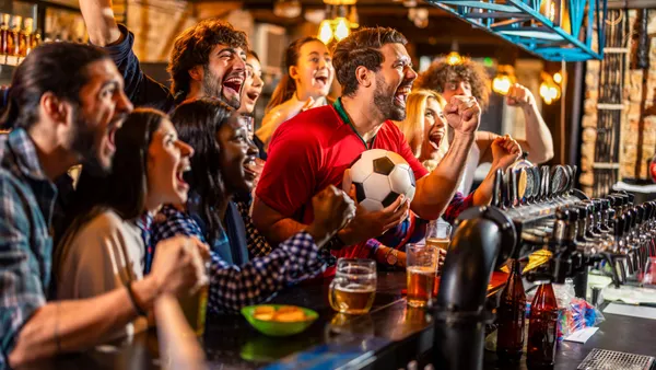 A group of people at a bar cheer while watching a soccer match. A person in the center is wearing red and holding a soccer ball.