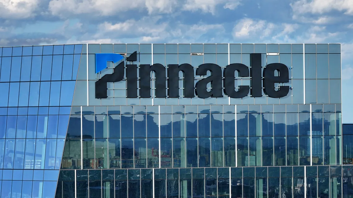 A glass office building with the word "Pinnacle" and its logo on the exterior is seen from high above Nashville's downtown.