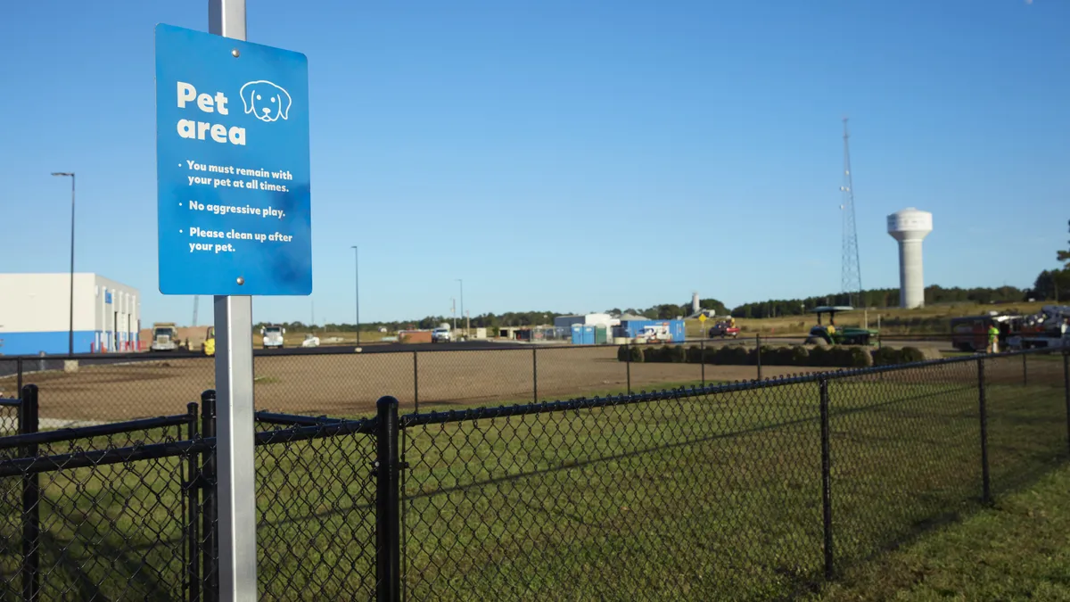 A photo of a grassy, fenced-in area. A blue and white sign with a picture of a dog's face on it reads "pet area. You must remain with your pet at all times. No aggressive play. Please clean up after your pet."