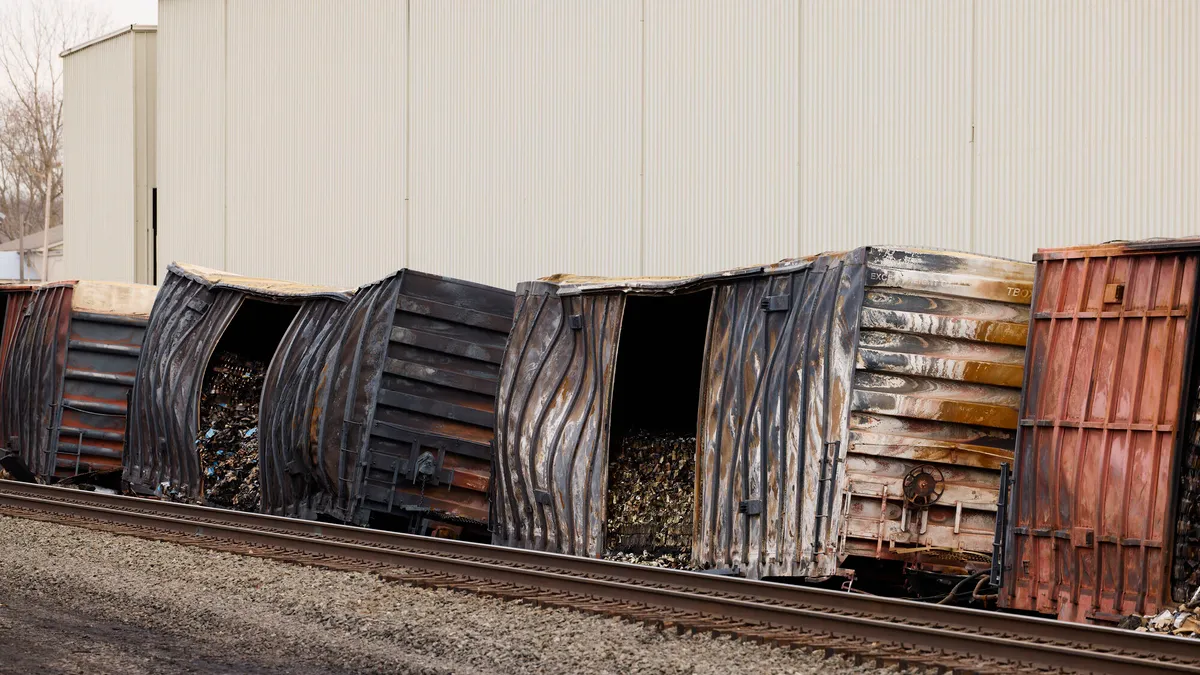 Several charred boxcars lie alongside a railroad track with a white industrial building behind them.